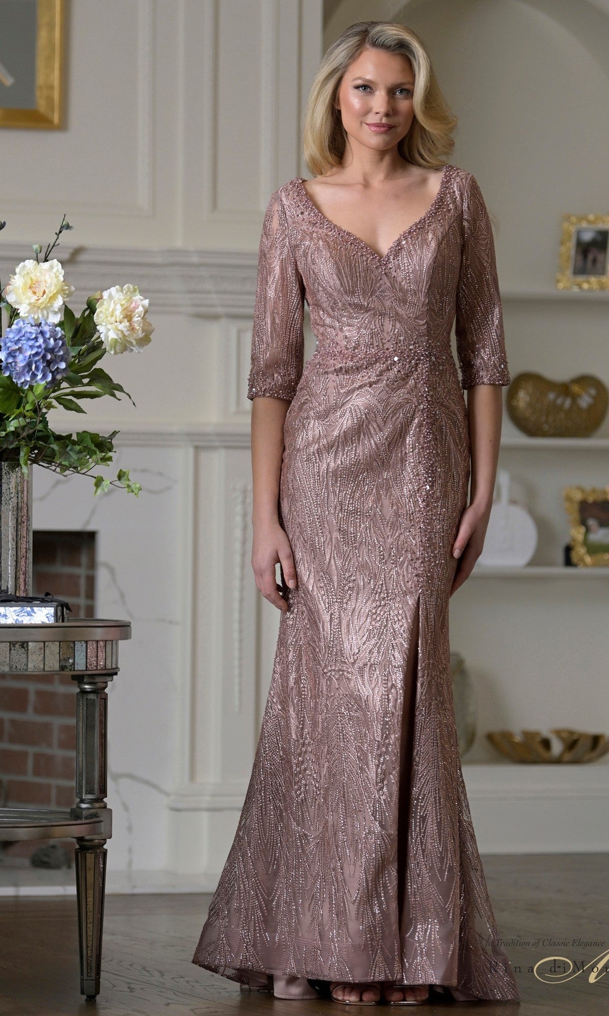 A woman stands indoors wearing the Rina Di Montella RD3530, a dark mauve, floor-length formal dress with three-quarter sleeves and a V-neckline; embroidered mesh details stand out amid stylish decor in the background.